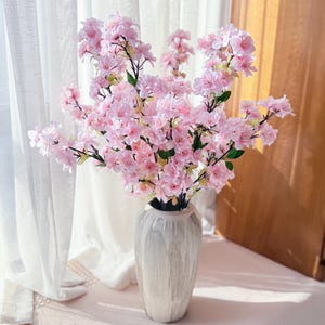 Op de afbeelding: Een boeket roze kunstbloemen in een getextureerde, gebroken witte vaas. De bloemen hebben meerdere lagen bloemblaadjes en groene stelen. De vaas is hoog en cilindrisch, met verticale groeven.