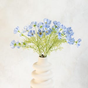 Könnte beinhalten: Ein dekoratives Arrangement aus hellblauen Blumen mit gelben Zentren und grünen Stielen in einer weißen, gestapelten Keramikvase. Die Vase steht auf einem weißen Sockel vor einem weißen Hintergrund. Die Blumen wirken zart.