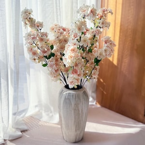 May include: A tall, textured, off-white vase filled with faux cherry blossom branches. The blossoms are a mix of white and pale pink, with green leaves. The vase is set against a sheer white curtain and a wooden door.