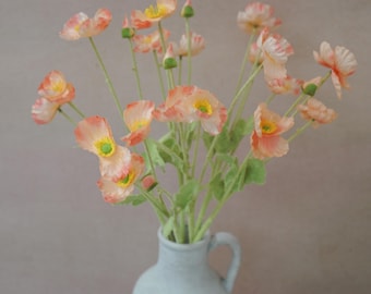 May include: A bouquet of peach-colored poppies with yellow centers and green stems in a light blue ceramic vase. The vase has a handle and sits on a yellow surface against a neutral background. The flowers are in full bloom.