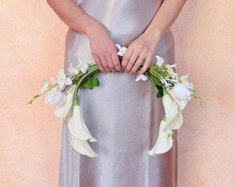 May include: A white floral wreath featuring calla lilies, roses, and other white flowers with green foliage. The wreath is held in front of a person wearing a silver dress. The background is a peach-colored wall.