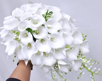 May include: A bridal bouquet of white calla lilies and lily of the valley. The bouquet features large, elegant calla lilies with a smooth texture, accented by small, delicate lily of the valley flowers and green foliage.