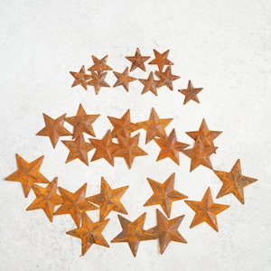 May include: A collection of rusty metal stars in various sizes arranged on a light gray surface. The stars have a weathered, orange-brown color, suggesting an aged or vintage aesthetic. The arrangement is scattered, creating a decorative element.
