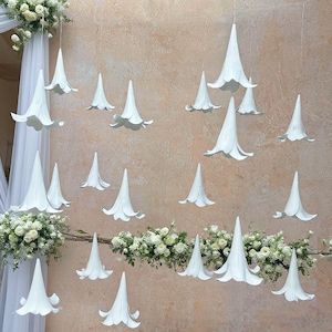 Puede incluir: Decoraciones florales blancas en forma de cono suspendidas del techo, intercaladas con arreglos florales. Las decoraciones están dispuestas en un patrón visualmente atractivo contra una pared de tono neutro, creando un ambiente elegante.