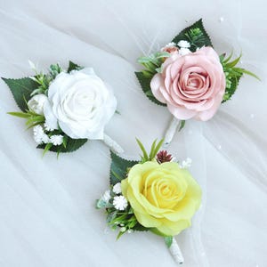 Puede incluir: Tres boutonnières artificiales de rosa con vegetación. Las rosas son blancas, rosas y amarillas.