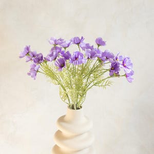 May include: A bouquet of purple flowers with green stems in a cream-colored, spiral-shaped vase. The vase sits on a white pedestal against a neutral background. The flowers have multiple petals and are in full bloom.