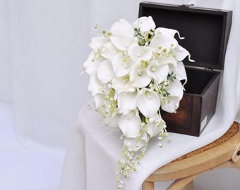 May include: A cascading bridal bouquet made of white calla lilies and greenery. The bouquet is arranged in a teardrop shape and is accented with pearl-like beads.