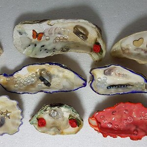 Könnte beinhalten: Eine Sammlung dekorativer Austernschalen, jede einzigartig verziert. Einige Schalen weisen kleine Ringe, Perlen und dekorative Elemente wie Schmetterlinge und Rosen auf. Die Schalen sind in verschiedenen Farben gehalten, darunter Weiß, Blau und Rot.