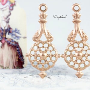 May include: Two gold-toned metal earring charms with a filigree design. The charms feature a circular centre with a floral pattern and a teardrop-shaped top.