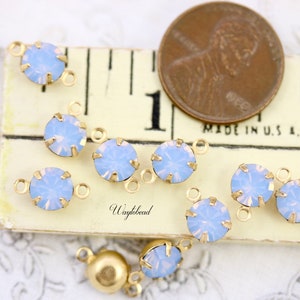 Puede incluir: Primer plano de un grupo de conectores de charms de &oacute;palo azul claro con engastes de metal dorado. Los charms est&aacute;n dispuestos sobre una superficie blanca con una regla y un centavo estadounidense para la escala. "Waylbead" est&aacute; impreso en la superficie.