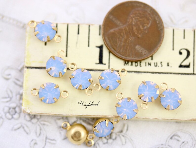Puede incluir: Primer plano de un grupo de conectores de charms de &oacute;palo azul claro con engastes de metal dorado. Los charms est&aacute;n dispuestos sobre una superficie blanca con una regla y un centavo estadounidense para la escala. "Waylbead" est&aacute; impreso en la superficie.