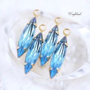 May include: Four blue crystal teardrop-shaped pendants with gold-colored metal accents. Each pendant has a loop at the top for hanging. The crystals have a faceted design, reflecting light. The pendants are arranged on a white surface.