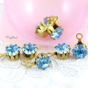 Puede incluir: Seis adornos de cristal azul claro con engastes y lazos de metal dorado. Los adornos están esparcidos sobre una superficie de encaje blanco.