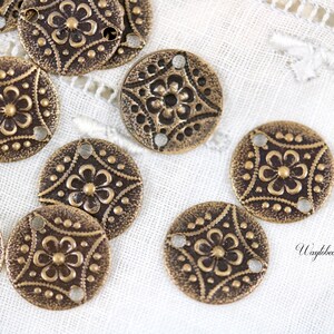 May include: A group of 10 antique gold-toned metal round charms with a floral design and three holes. The charms are on a white fabric background.