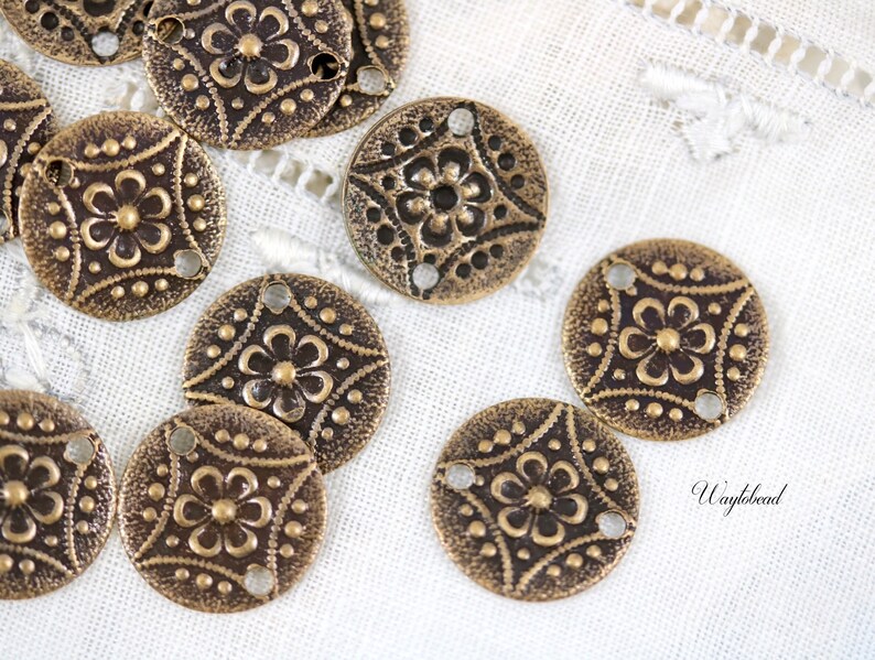 May include: A group of 10 antique gold-toned metal round charms with a floral design and three holes. The charms are on a white fabric background.