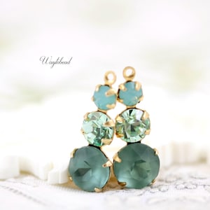 May include: Dangle earrings with three graduated, round, faceted gemstones in teal and green, set in gold-coloured metal. The earrings have a post and loop design. The word "Waylbead" is written in cursive on the left side.
