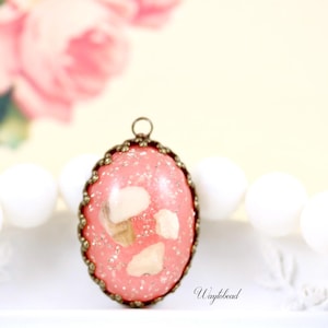 May include: A pink and white oval pendant with a gold-tone metal frame. The pendant has a glittery finish and features a white design inside.