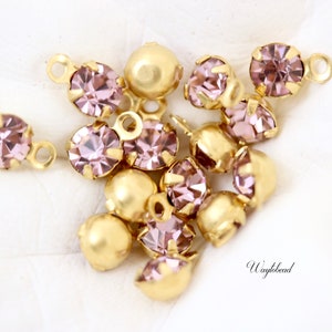 Puede incluir: Un grupo de pequeños diamantes de imitación de vidrio rosa facetados con engastes y lazos dorados. Los diamantes de imitación están esparcidos sobre una superficie blanca.