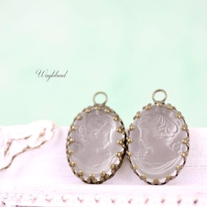 May include: Two oval-shaped cameo pendants with a woman's profile etched in the center. The pendants have a clear, frosted appearance and are framed with a decorative, antique gold-colored metal. Each pendant has a small loop at the top.
