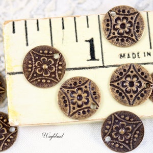 May include: A collection of 10 antique brass charms with a floral design. Each charm has two holes and is approximately 1 inch in diameter. The charms are arranged on a white surface with a ruler in the background.
