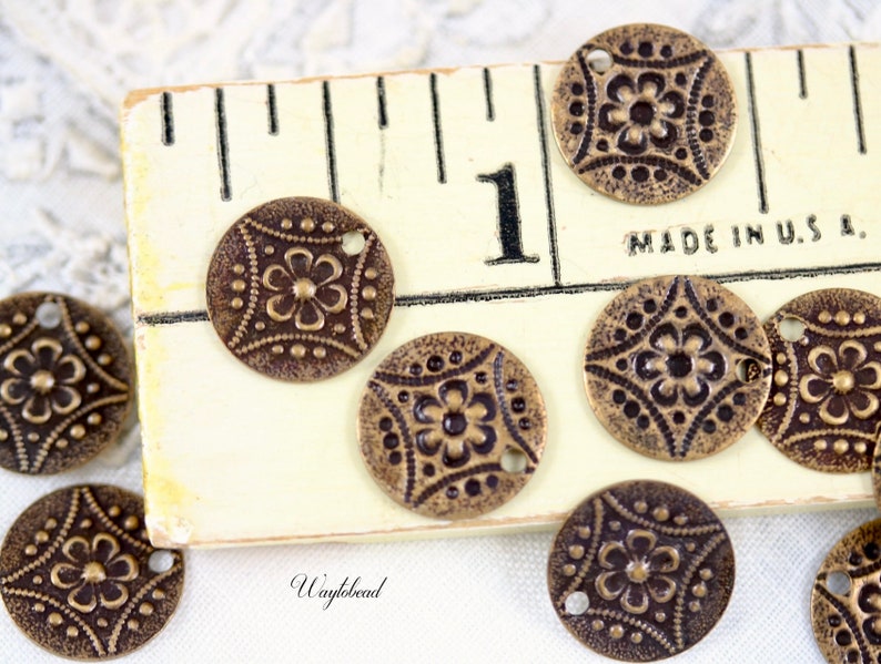 May include: A collection of 10 antique brass charms with a floral design. Each charm has two holes and is approximately 1 inch in diameter. The charms are arranged on a white surface with a ruler in the background.