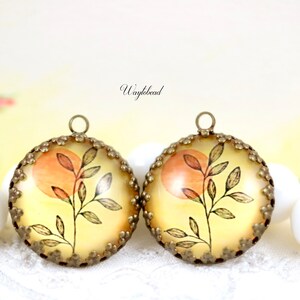 Puede incluir: Dos abalorios redondos de metal dorado con un diseño floral. Cada abalorio presenta una rama delicada con hojas y un círculo de color melocotón en el centro. Los abalorios tienen un borde festoneado.
