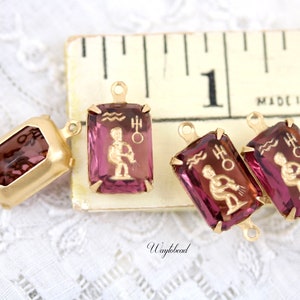 May include: Three gold-tone metal charms with purple glass insets featuring the astrological symbol for Aquarius. The charms have a loop at the top for attaching to a necklace or bracelet. The charms are displayed on a ruler with the text "Made in U.S.A."