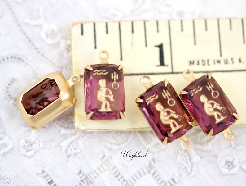 May include: Three gold-tone metal charms with purple glass insets featuring the astrological symbol for Aquarius. The charms have a loop at the top for attaching to a necklace or bracelet. The charms are displayed on a ruler with the text "Made in U.S.A."