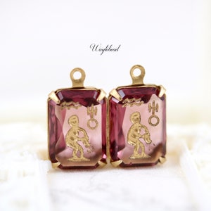 May include: A pair of gold-tone earrings with pink glass stones featuring the astrological symbol for Aquarius.