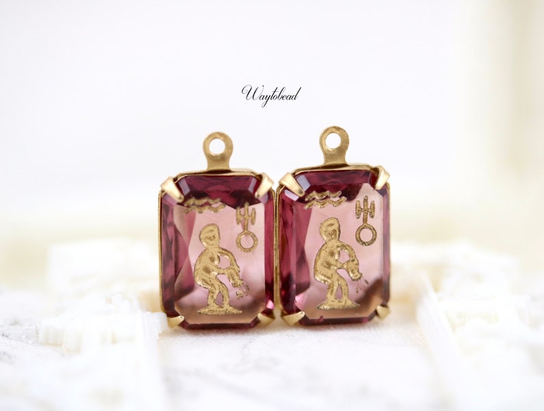May include: A pair of gold-tone earrings with pink glass stones featuring the astrological symbol for Aquarius.