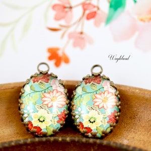 May include: Two oval-shaped, antique bronze, floral patterned glass cabochons with a metal loop at the top. The cabochons have a light blue background with red, pink, yellow, and green floral designs.