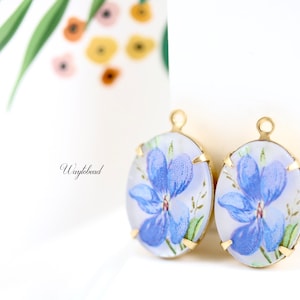 Può includere: Due ciondoli ovali in metallo dorato con un design floreale blu. I ciondoli hanno un anello nella parte superiore per essere attaccati a gioielli.