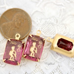 May include: Two gold-toned pendant charms with pink glass insets featuring the astrological symbol for Aquarius. The charms have a loop at the top for attaching to a necklace or bracelet.