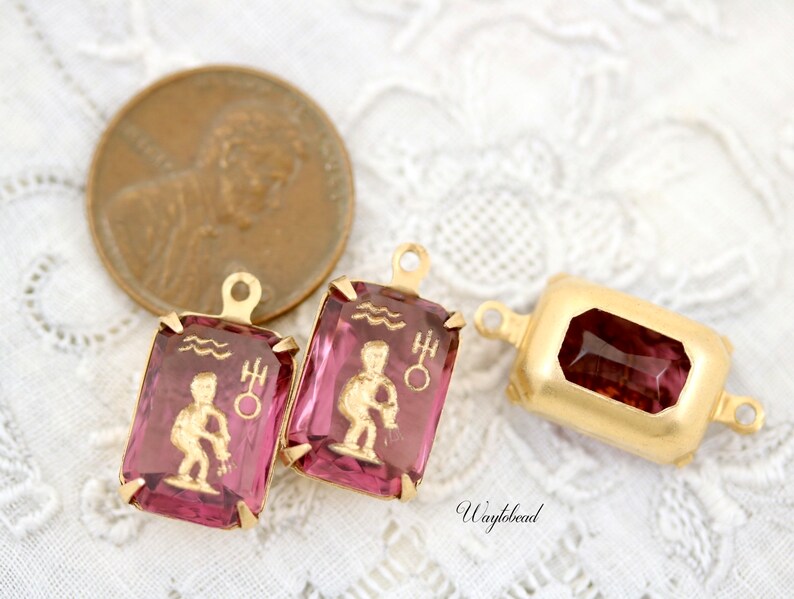 May include: Two gold-toned pendant charms with pink glass insets featuring the astrological symbol for Aquarius. The charms have a loop at the top for attaching to a necklace or bracelet.