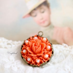 May include: A coral-colored rose pendant with white pearl accents, set in a bronze-toned metal frame. The pendant is on a white lace background.