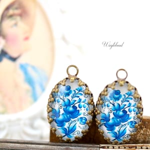 May include: Two oval-shaped glass charms with a blue and white floral design. The charms have a gold-toned metal frame with a beaded edge.