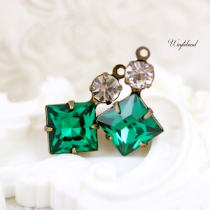 Op de afbeelding: Een close-up van een groene en heldere kristallen hanger met een messing setting. De hanger heeft twee vierkante groene kristallen en één ronde heldere kristal.