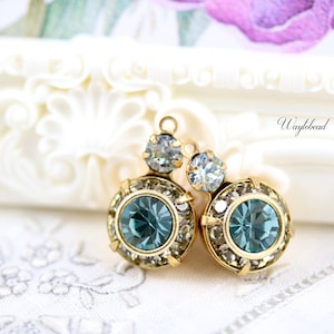 May include: A pair of gold-toned earrings with teal and clear crystal accents. Each earring features a large teal center stone surrounded by smaller clear crystals. The earrings have a vintage style and are set against a white background.
