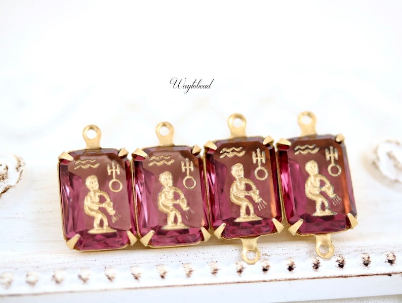 May include: Four gold-tone charms with pink glass insets featuring the astrological sign Aquarius. Each charm has a loop at the top for attaching to a necklace or bracelet.