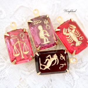 May include: Four gold-tone charms featuring zodiac signs. The charms are rectangular with a clear glass centre. The signs are Libra, Aquarius, Cancer, and Capricorn. The charms are pink, red, and burgundy.