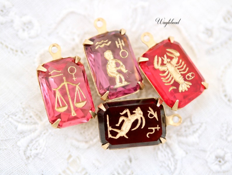 May include: Four gold-tone charms featuring zodiac signs. The charms are rectangular with a clear glass centre. The signs are Libra, Aquarius, Cancer, and Capricorn. The charms are pink, red, and burgundy.