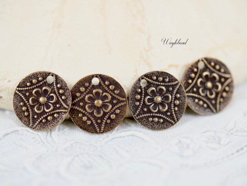 May include: Four antique brass-colored metal charms with a floral design and a hole for hanging. The charms are round and have a textured surface.