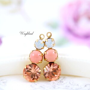 May include: A pair of dangling earrings with three tiers of colorful stones. The top stone is a light blue, the middle is a soft pink, and the bottom is a peach color. The earrings have a gold-colored metal setting. The word "Waylbead" is written in cursive.