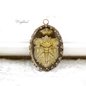 May include: An oval pendant with a brown background and a gold emblem featuring a crown and a shield design. The pendant is framed with small, gold-colored beads and has a loop at the top. The text "Waytobead" is in the upper left corner.