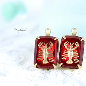 May include: Two red glass charms with gold lobster designs. The charms have a gold loop at the top for attaching to jewelry.