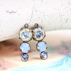 Può includere: Primo piano di due orecchini con cristalli azzurri e trasparenti. Gli orecchini hanno uno stile vintage con una montatura in metallo dorato. Il testo "Waylbead" è visibile.