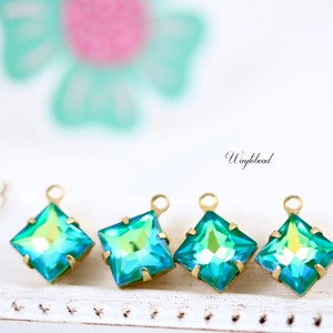 May include: Four square-shaped turquoise iridescent glass charms with gold jump rings. The charms are on a white surface.