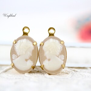 May include: Two oval-shaped cameo pendants with gold-tone settings. Each pendant features a white cameo of a woman's profile.