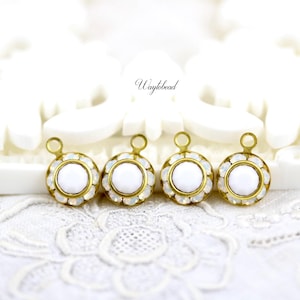 Op de afbeelding: Vier goudkleurige metalen bedels met witte ronde cabochons en heldere kristallen accenten. De bedels hebben een lus aan de bovenkant om aan sieraden of andere knutselwerkjes te bevestigen.