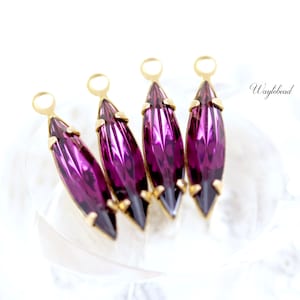 May include: Four purple marquise-shaped crystal charms with gold-toned jump rings. The charms are arranged in a row on a white surface.